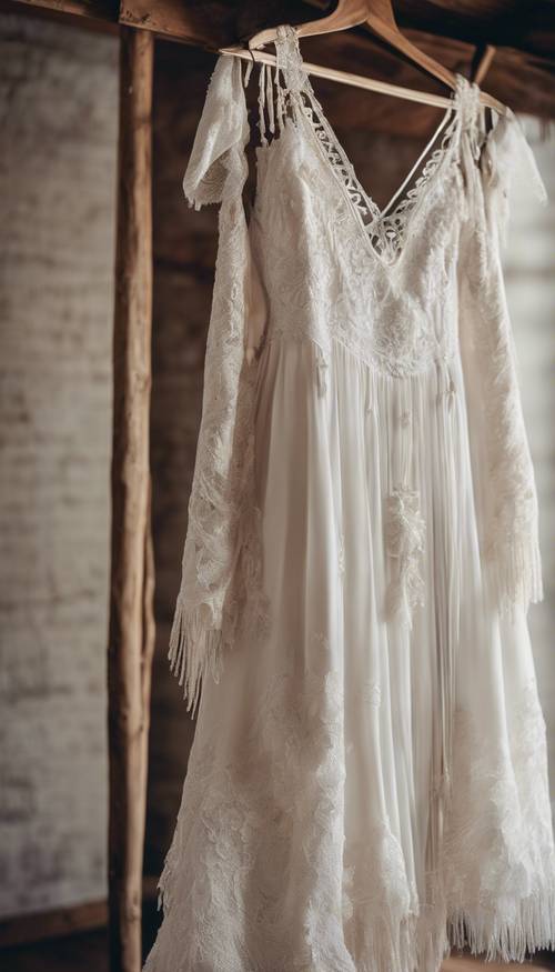 Um vestido de noiva Boho branco com detalhes em renda e borlas, pendurado em um cabide de madeira vintage.