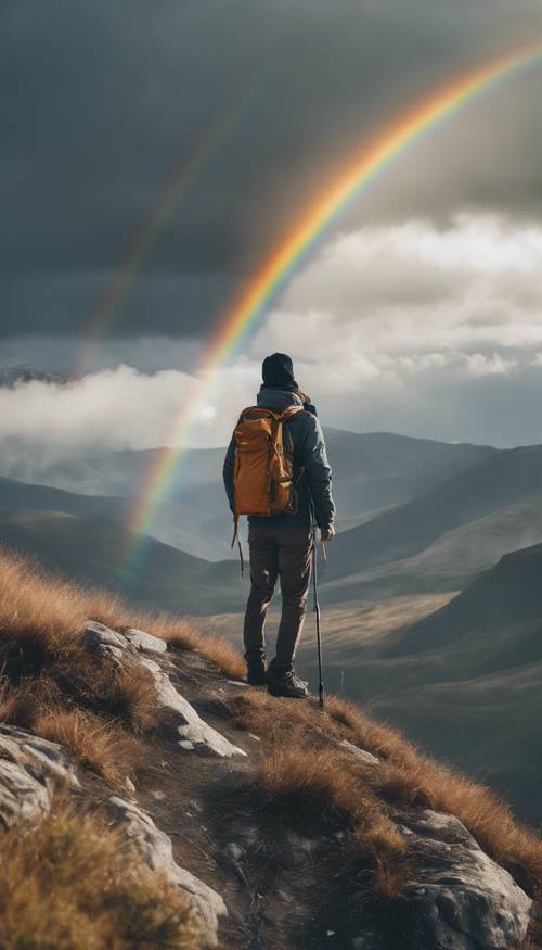 Un randonneur debout au sommet d’une montagne contemplant un arc-en-ciel noir.