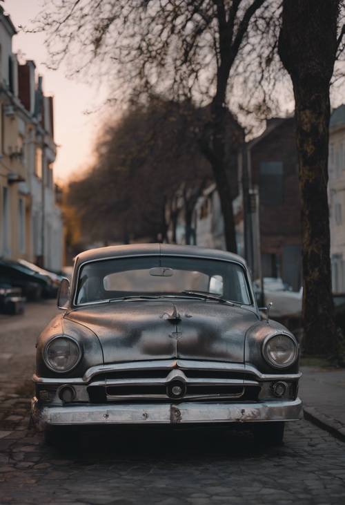 Sebuah mobil antik usang dengan cat abu-abu metalik diparkir di jalan yang sepi saat senja.