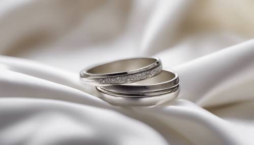 Silver wedding bands resting on a white satin cushion. Tapet [5b20436196064a1c869b]