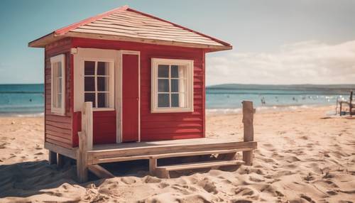 A beige and red wooden beach hut on a sandy beach on a bright sunny day.