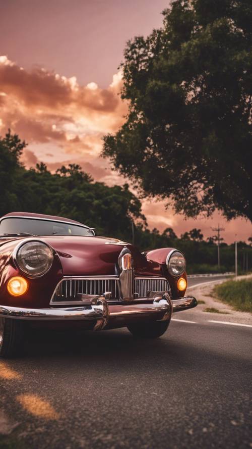 Mobil vintage merah marun yang keren melaju di jalan raya saat matahari terbenam.