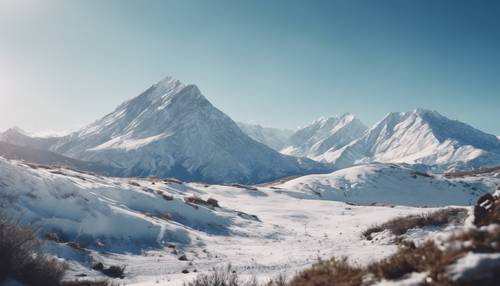 Berrak mavi gökyüzünün altında karla kaplı dağ silsilesinin sakin manzarası.