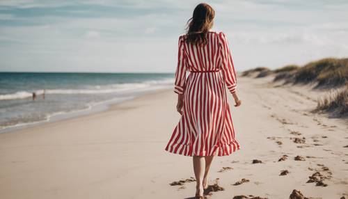 A stylish woman wearing a red and white striped dress walking alongside a serene beach.