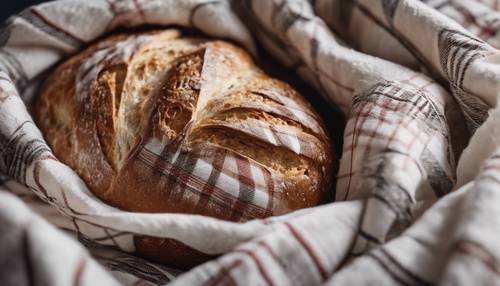 A fresh loaf of artisanal sourdough bread wrapped in a white plaid dishcloth. Tapet [49711ac0ab2548f0a972]