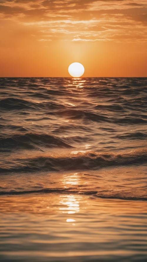 Pomarańczowe słońce zachodzące nad spokojnym oceanem, odbijające na wodzie jego bogate i ciepłe kolory.