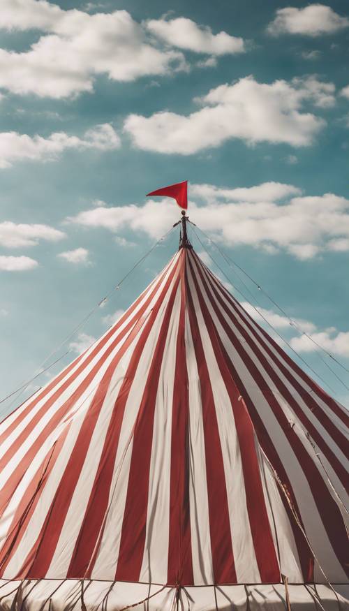 Tenda sirkus antik bergaris merah dan putih menghadap langit biru dengan awan tipis mengambang.