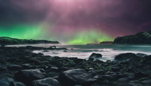 A misty panorama of a black rocky shoreline being met with iridescent, green northern lights.