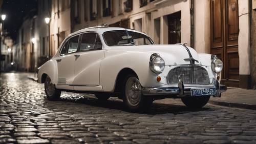 White vintage car, with a polished silver trim, parked in a moonlit cobbled street.
