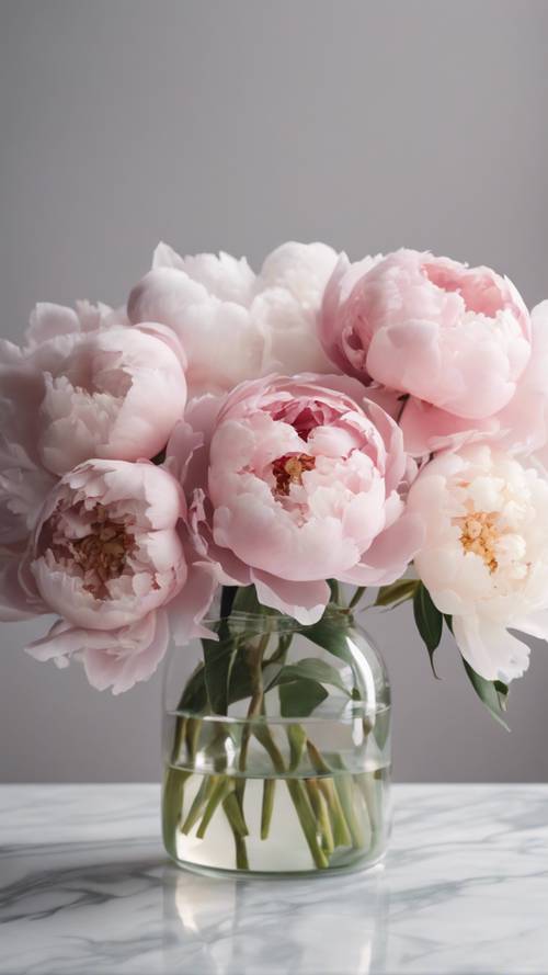 A bunch of stylish peonies in pastel colors arranged artistically in a clear glass vase on the white marble table.